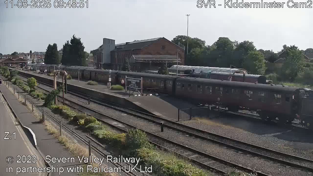 A long train of dark-colored passenger cars sits at a station on a sunny day.