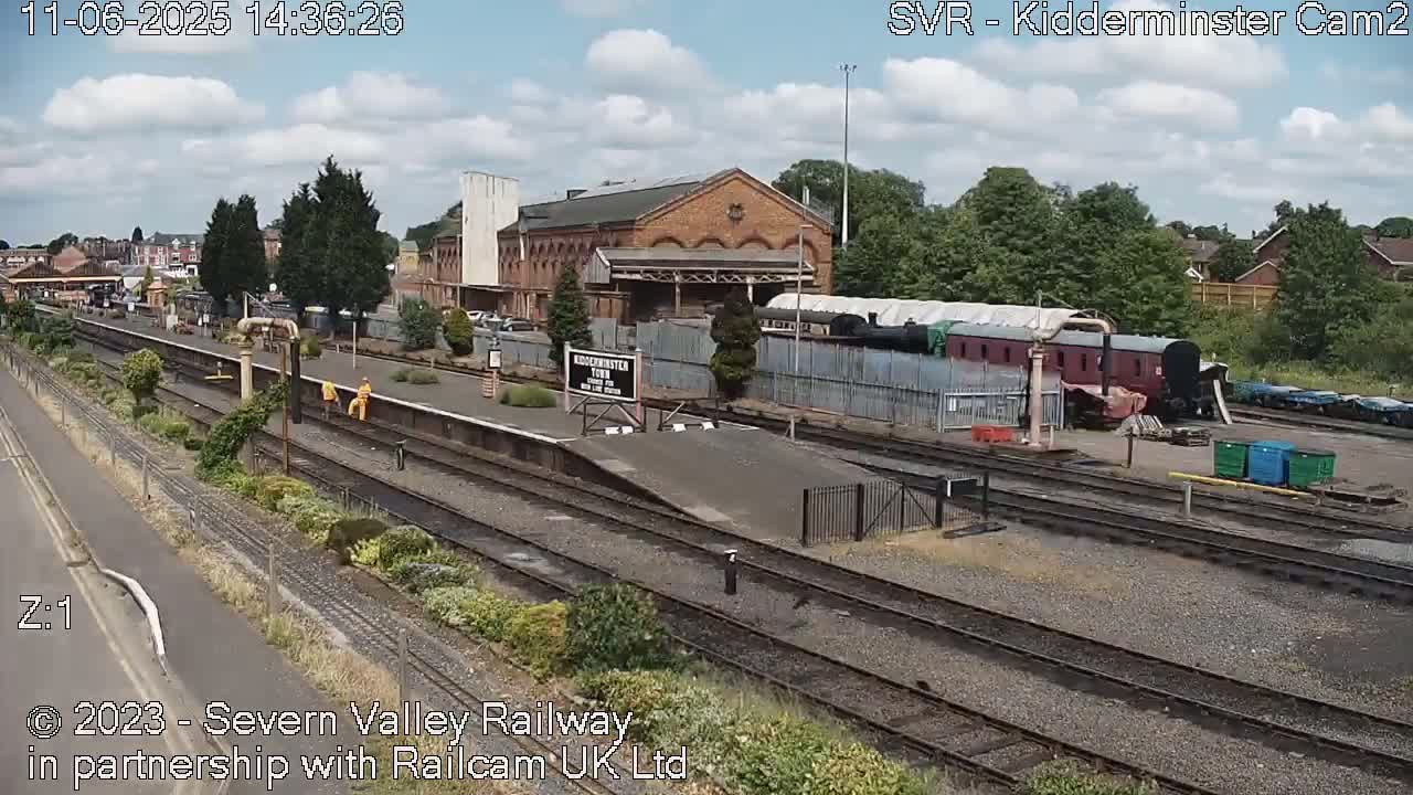 A partly cloudy daytime view of a train station with multiple train tracks, a brick building, and several railway cars.