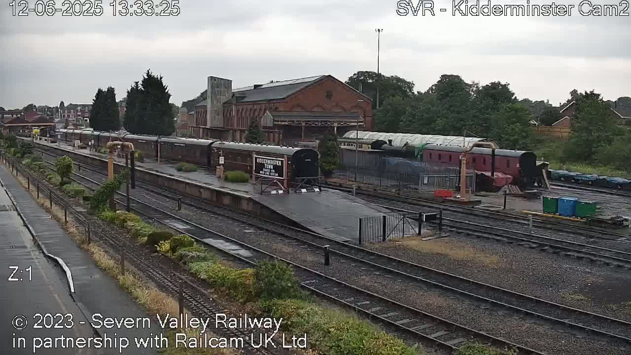 A train station on an overcast day with several dark-colored passenger cars on the tracks and a large brick building in the background.