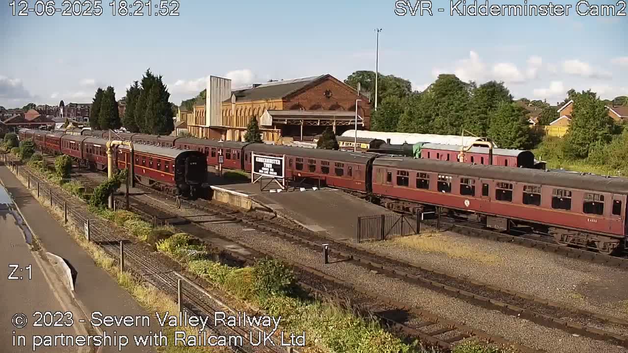 A long maroon passenger train sits at a station on a sunny day, next to a brick building.