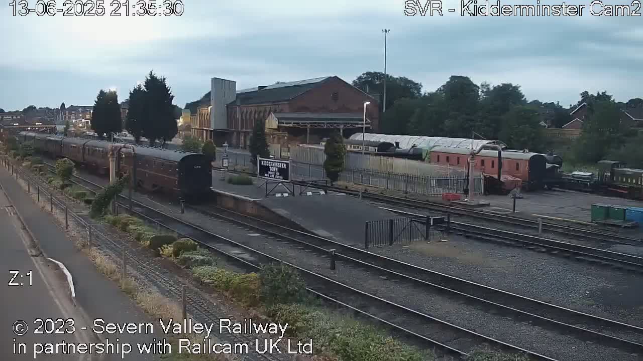 Under an overcast sky, a long train of dark brown passenger cars sits on the tracks next to a brick train station and other railway cars.