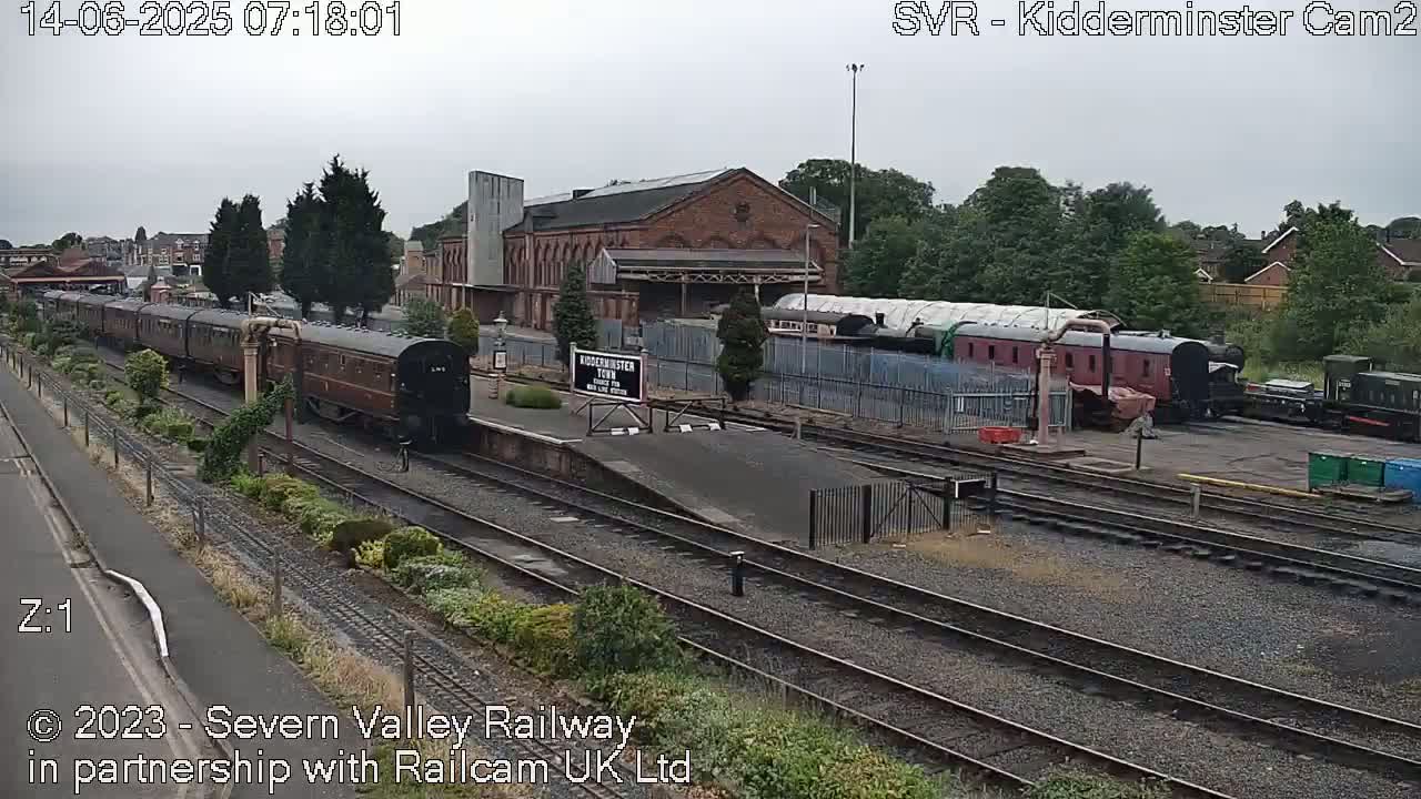 Under an overcast sky, a long train of dark brown passenger cars sits on the tracks next to a brick train station and other railway cars.