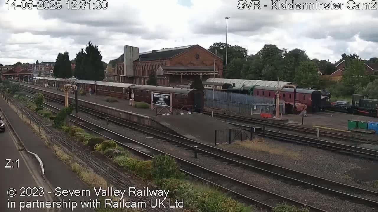 Under a partly cloudy sky, a railway station with several train cars and a large brick building is shown.