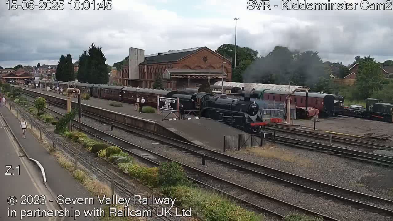 A steam train is pulling into a train station on an overcast day.