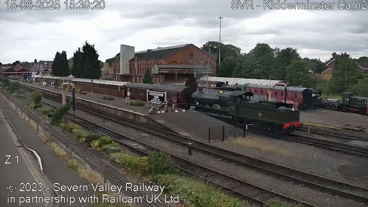 A steam train sits at a railway station on an overcast day, with several passenger cars and other trains visible nearby.