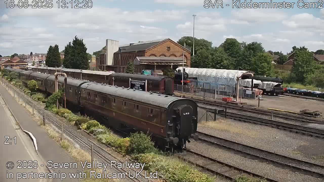 A long train of dark passenger cars is on a railway track next to a brick building, under a partly cloudy sky.