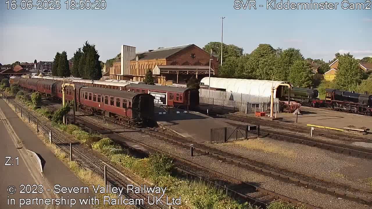 A sunny day at a train station shows multiple railway cars and locomotives parked on tracks beside a brick building and other structures.