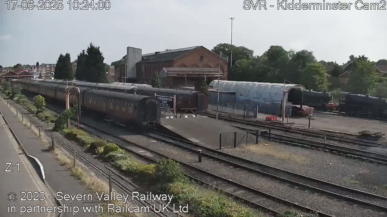 A long train of dark passenger cars sits on a railway track next to a brick building on a partly sunny day.