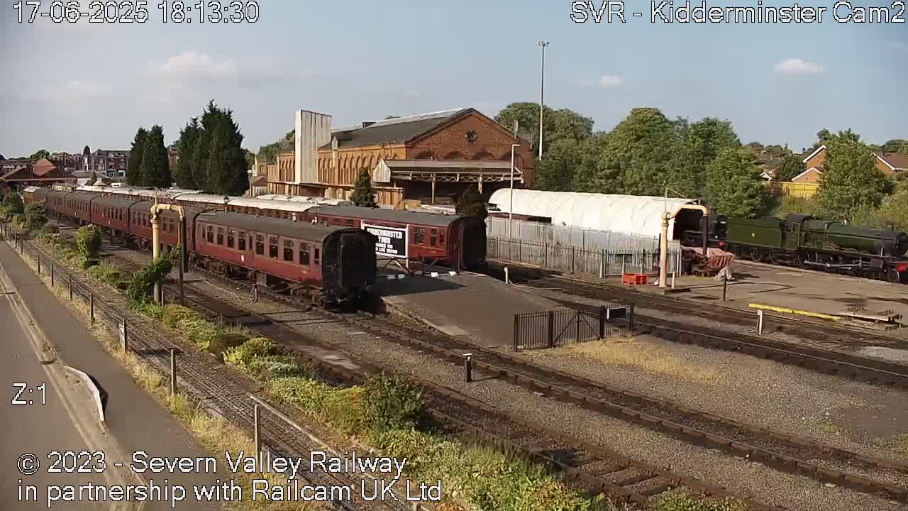 A long train of maroon passenger cars sits on a railway track next to a brick building and a dark green locomotive under a mostly sunny sky.