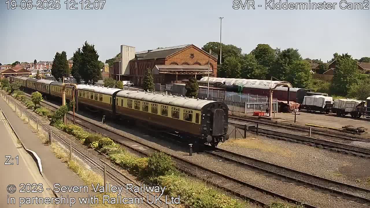 A long cream and brown passenger train moves past a brick train station on a sunny day.
