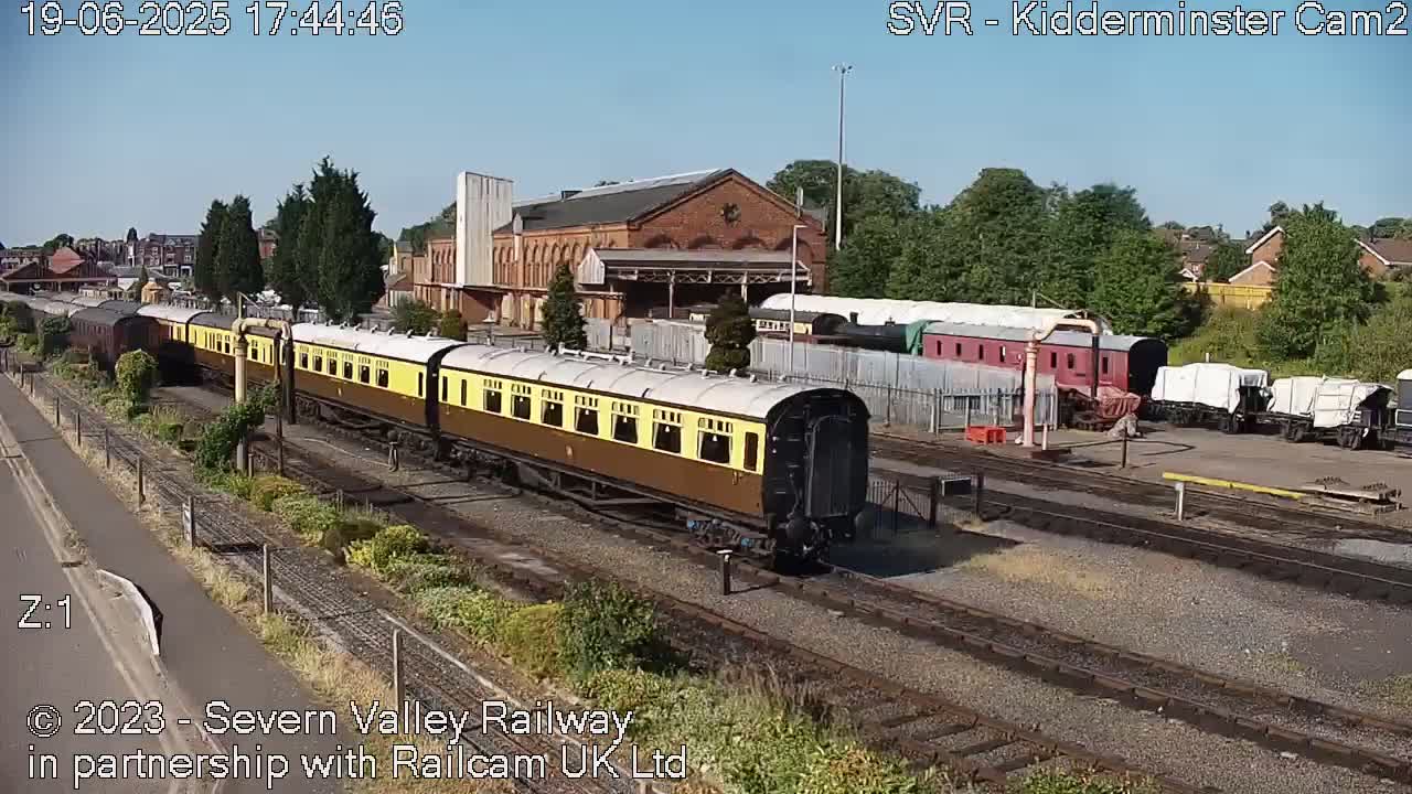 A long train of brown and yellow passenger cars sits on a railway track next to a brick building under a sunny sky.
