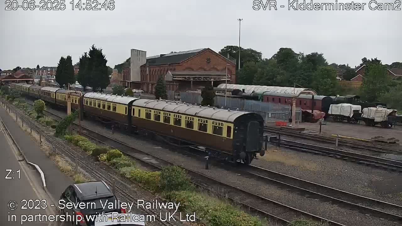 A long passenger train of dark brown and tan cars sits on railroad tracks next to a brick building under an overcast sky.