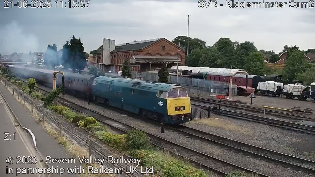 A teal and yellow diesel locomotive pulls a long train past a brick railway station on an overcast day.