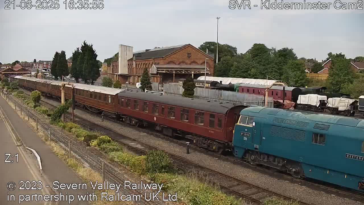 A teal diesel locomotive is coupled to a long string of maroon passenger cars on a railway track under an overcast sky.