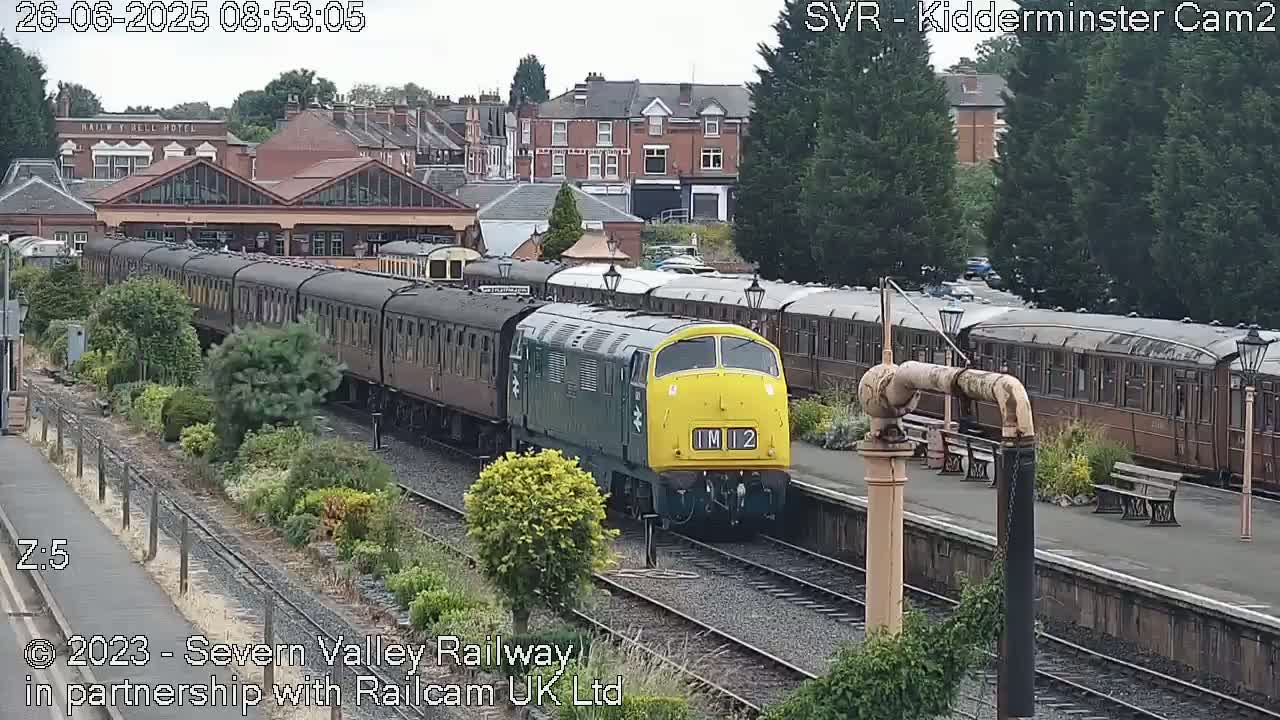 A green and yellow diesel locomotive pulls a long train of brown passenger cars past a station on an overcast day.