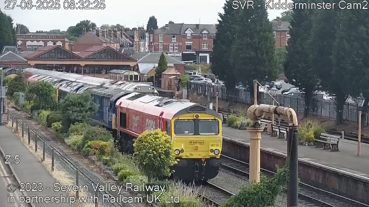 A long freight train, led by a red and yellow locomotive, is passing through a train station on a partly sunny day.