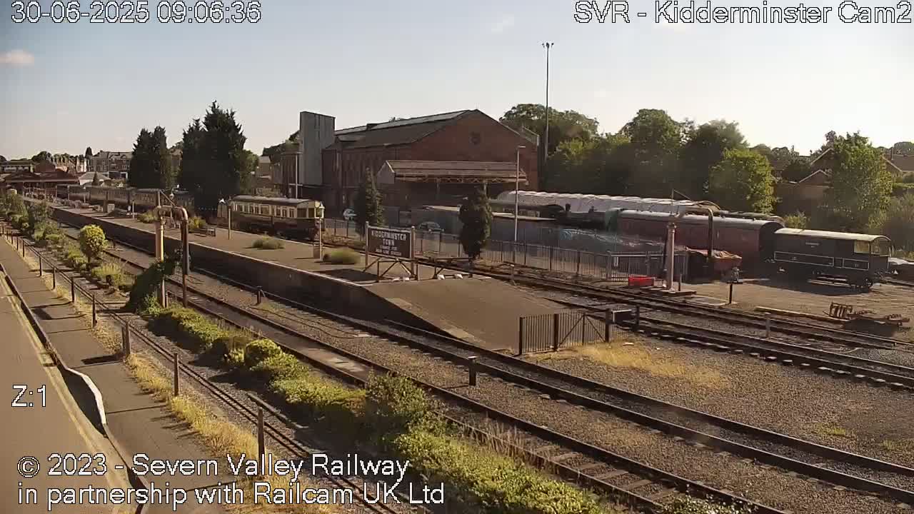 A sunny day at a train station with multiple railway cars and tracks, some of which are parked near a brick building.