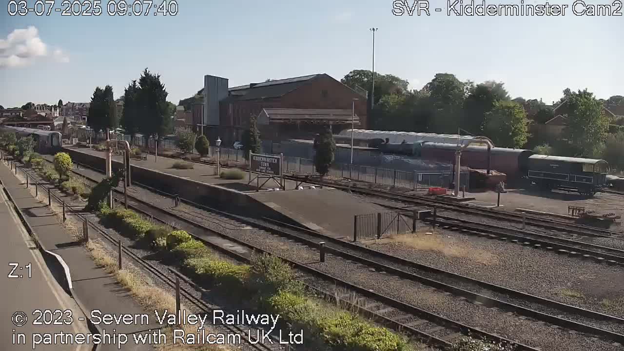 A sunny day at a train station shows a passenger train and several railcars parked on the tracks, with buildings and trees in the background.