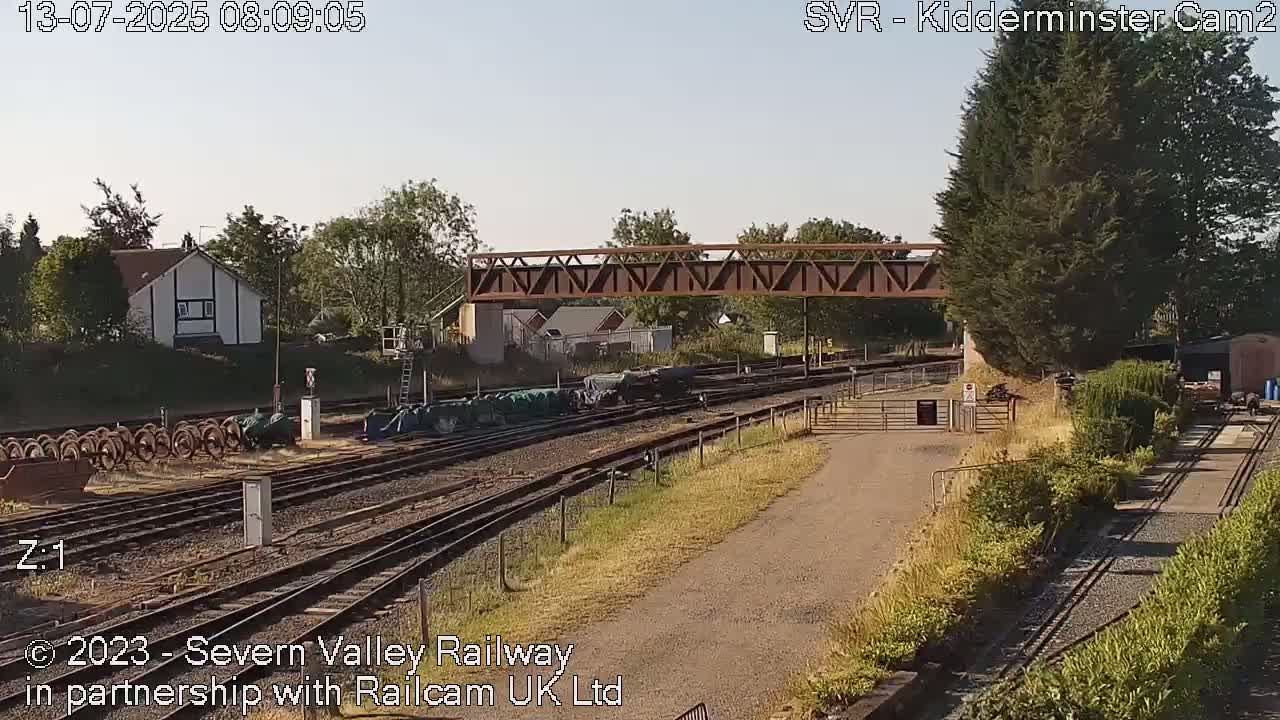 A sunny day at a railway yard shows train cars parked on multiple tracks near a pedestrian bridge and a small building.