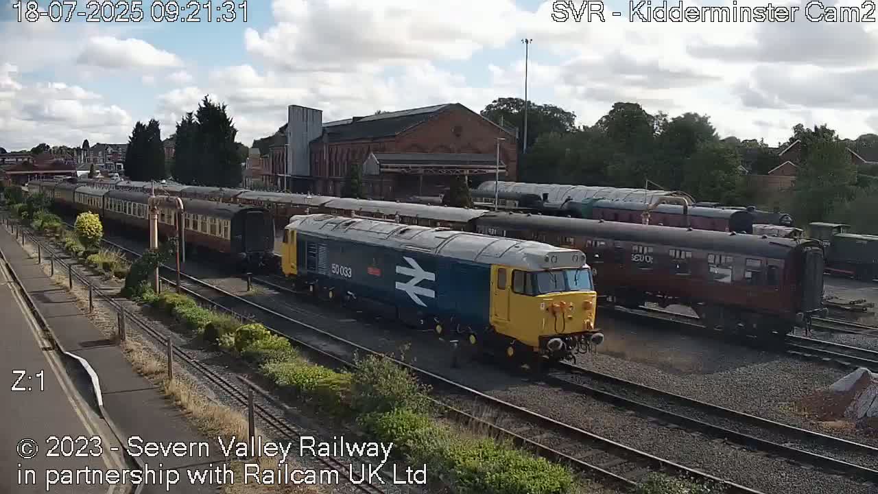 A blue and yellow diesel locomotive leads a train of maroon passenger cars on a railway line under a partly cloudy sky.