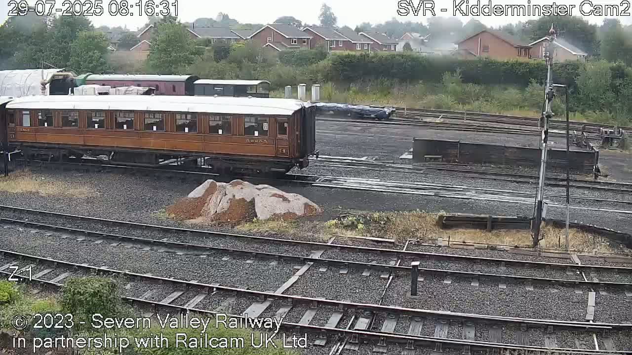 A brown wooden passenger railcar sits on train tracks in a rail yard on an overcast day.