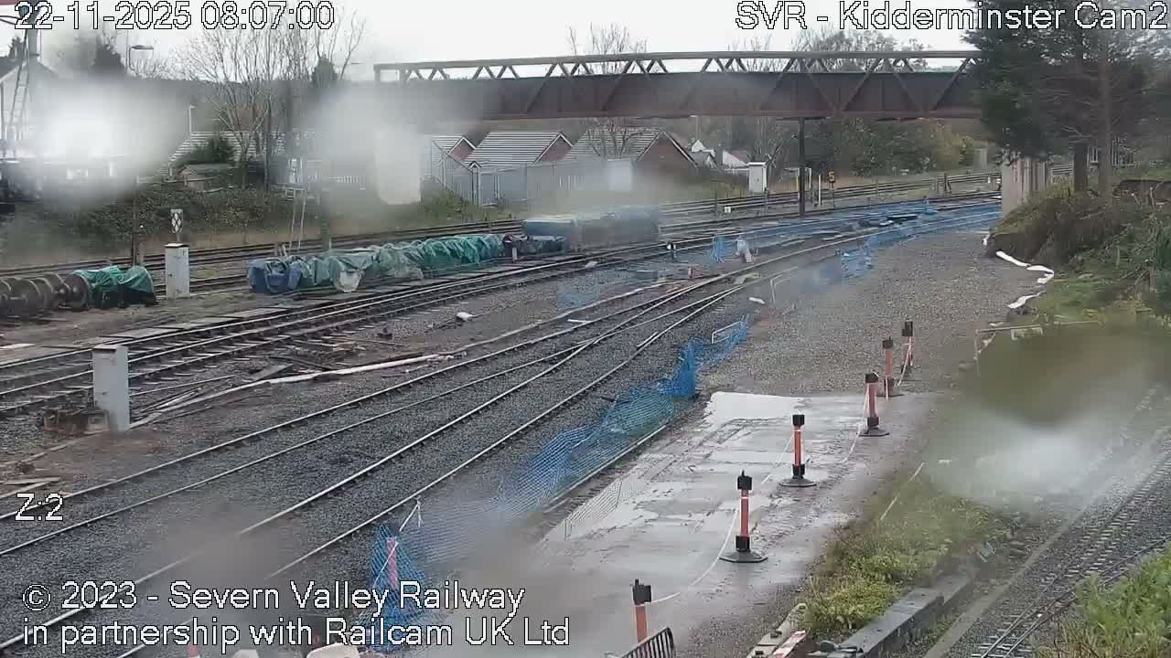 A misty, overcast view of a railway yard features multiple tracks, covered equipment, safety netting, and an overhead metal truss bridge.