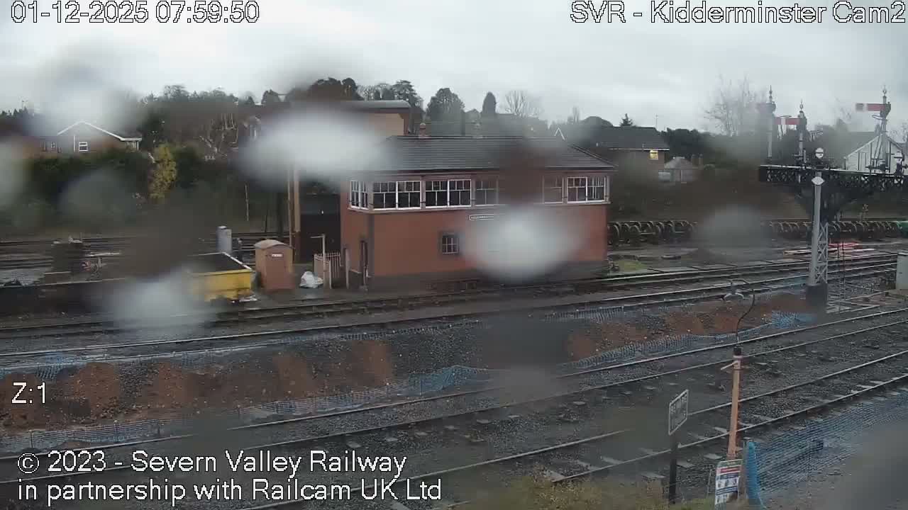 A brick railway signal box stands beside multiple tracks and a signal gantry under a grey, overcast, and rainy sky.