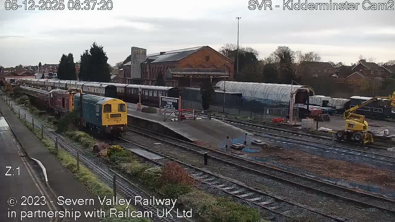 Under an overcast sky, a train station and railway yard are visible, featuring a yellow and blue locomotive on tracks beside a damp platform, multiple train carriages, a large brick building, various rail equipment including a yellow track-laying machine, and distant residential homes.