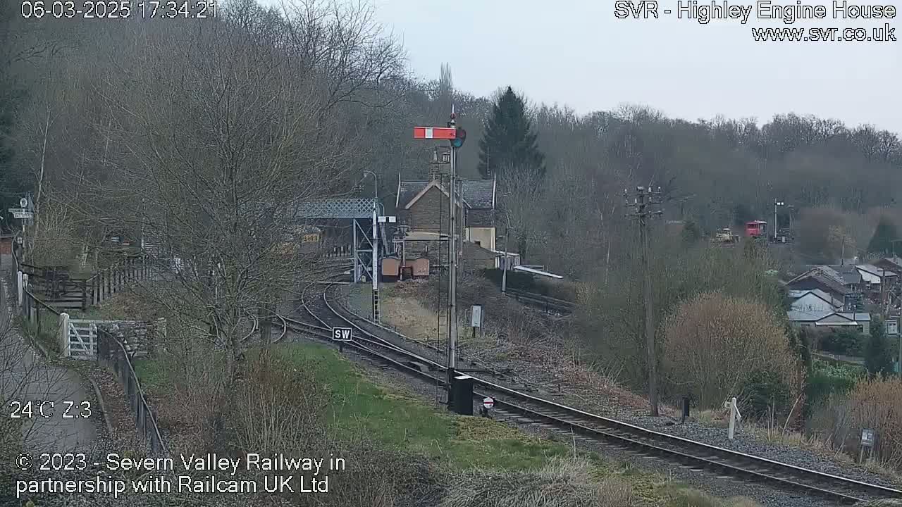 A railway station with train tracks and signals is seen on an overcast day, partially surrounded by trees and buildings.