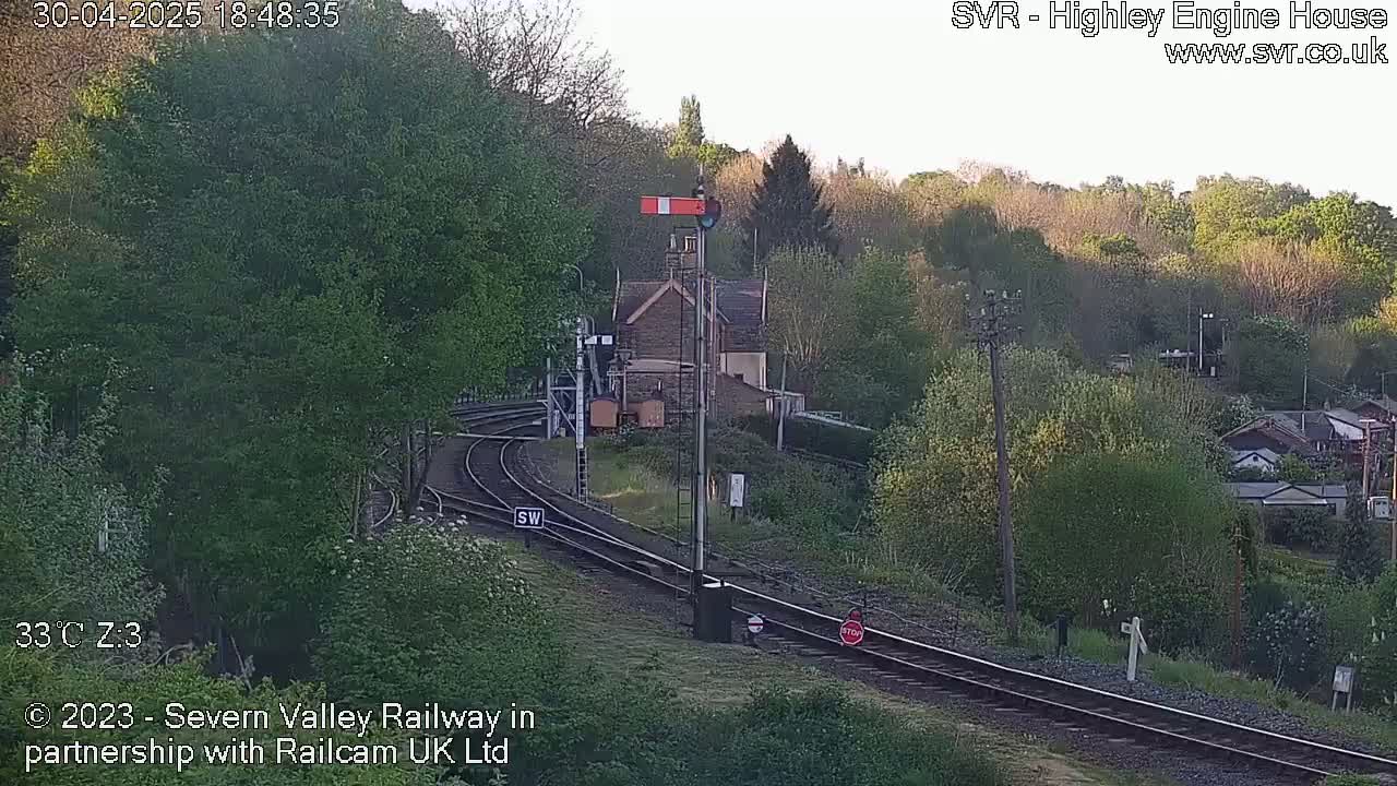 A railway line curves through a hilly landscape under a clear, bright sky, with a small stone building and lush green vegetation visible.