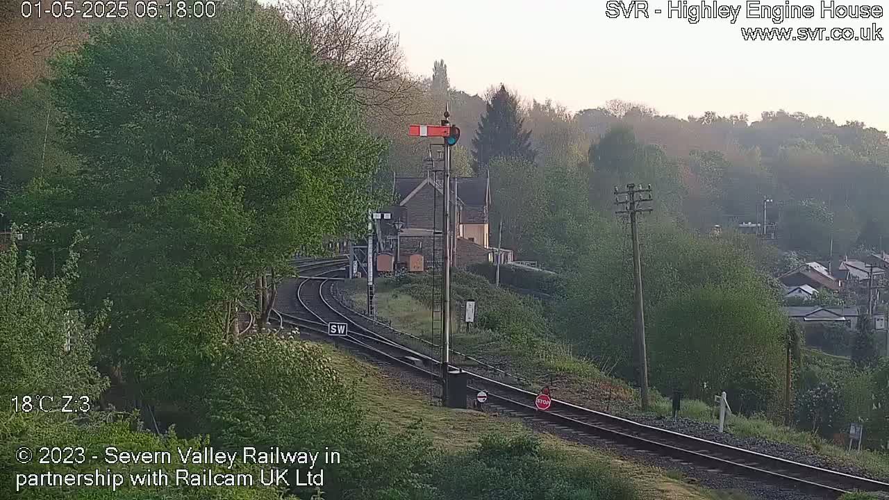 A hazy sunrise illuminates a railway scene with curving tracks, lush greenery, and a signal tower near a stone building.