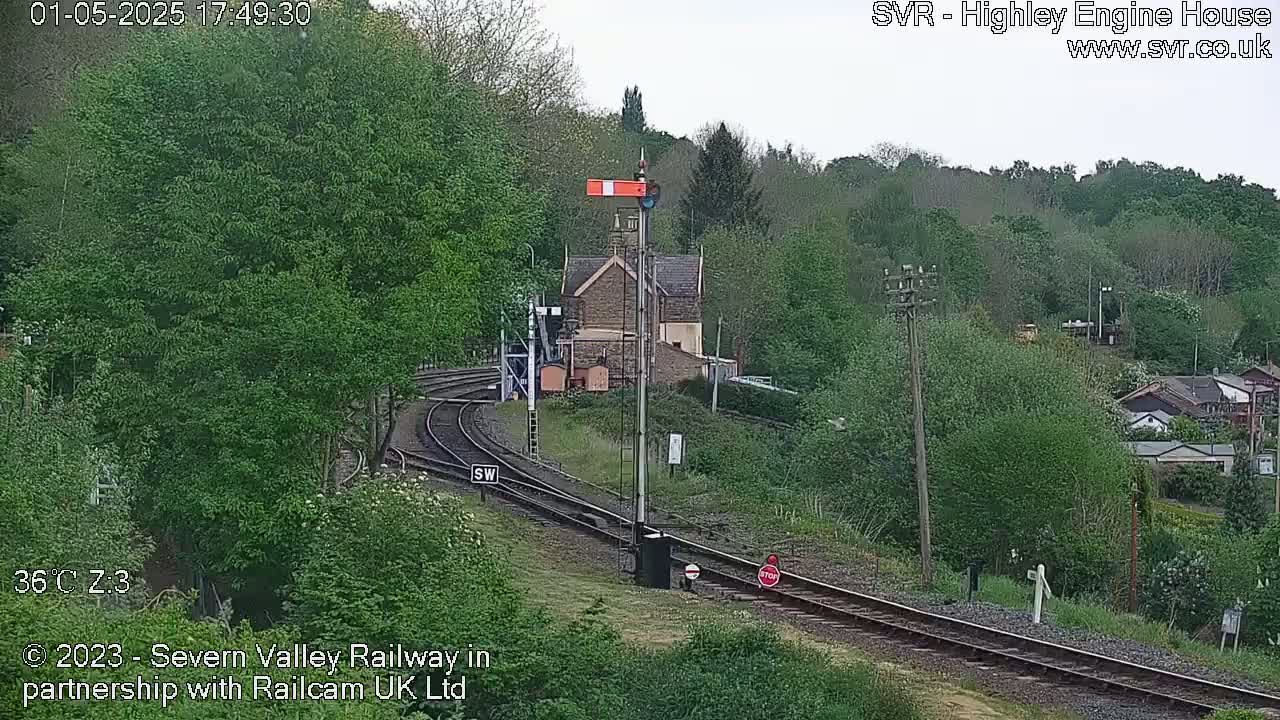 A railway line curves through a green, leafy landscape past a stone building under a partly cloudy sky.