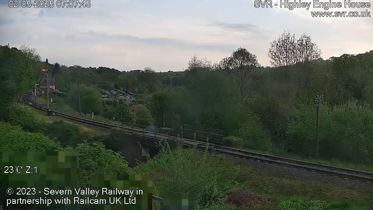 A train travels along a railway track that curves through a lush green valley under a partly cloudy sky.