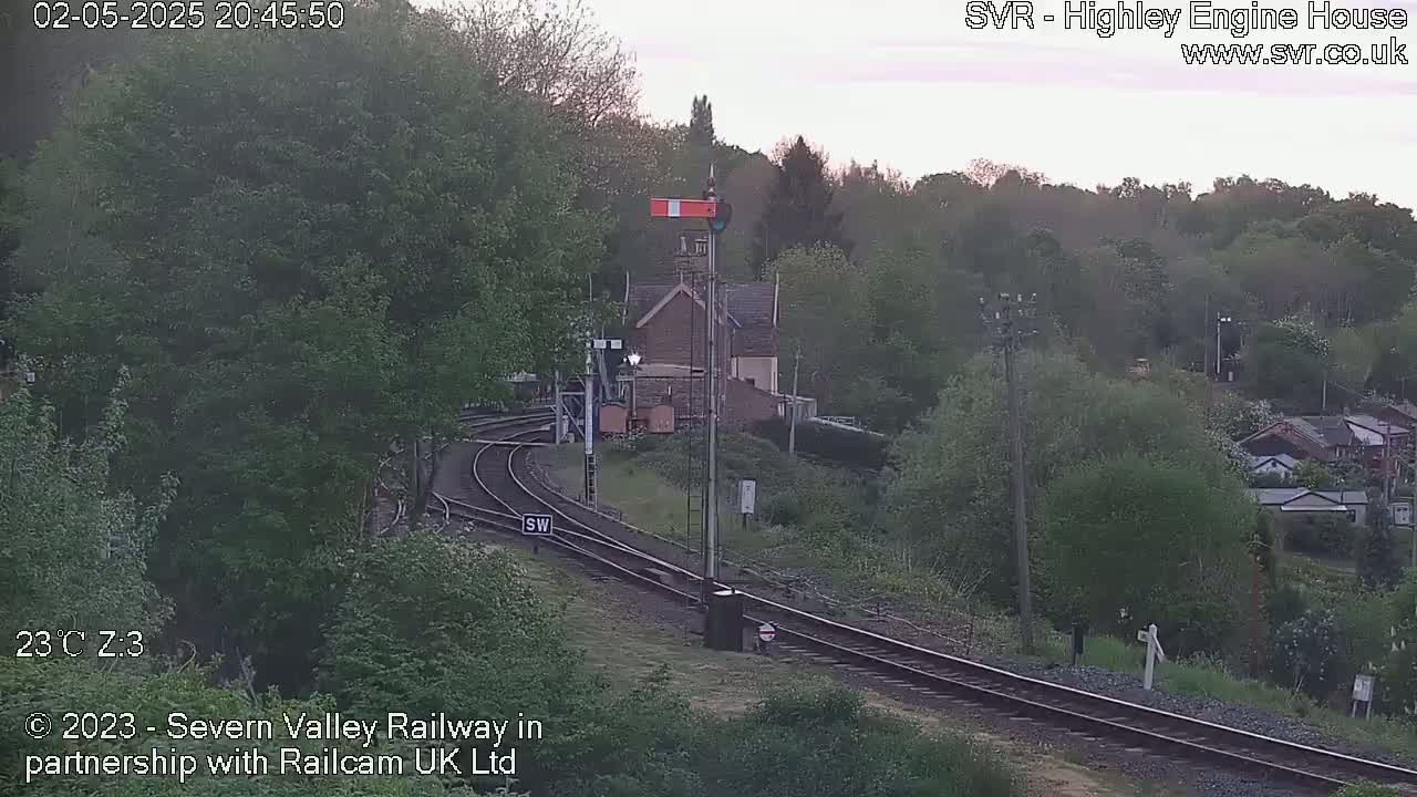 A railway line curves through a green, wooded area under a twilight sky, passing near a small building.