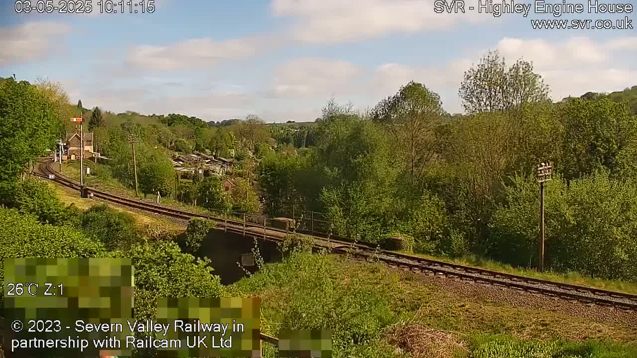 A sunny day reveals a railway track winding through a verdant valley, passing a small station and under a bridge, with lush green trees and foliage lining the landscape.