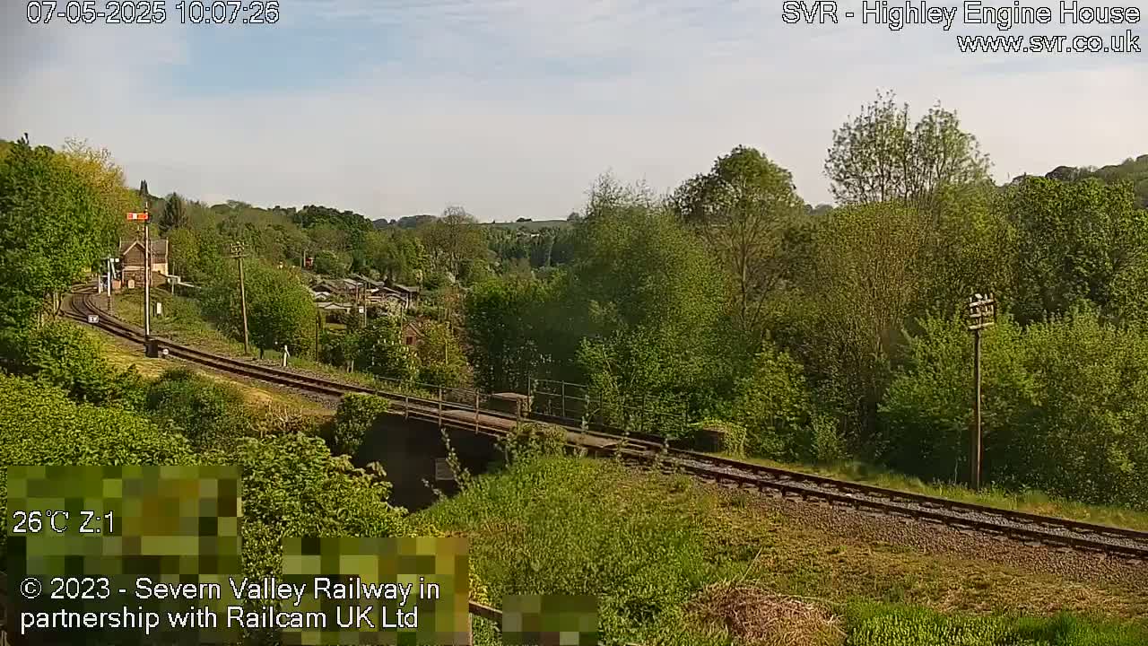 A railway track curves gently through a lush green landscape under a clear sky, passing over a small bridge.