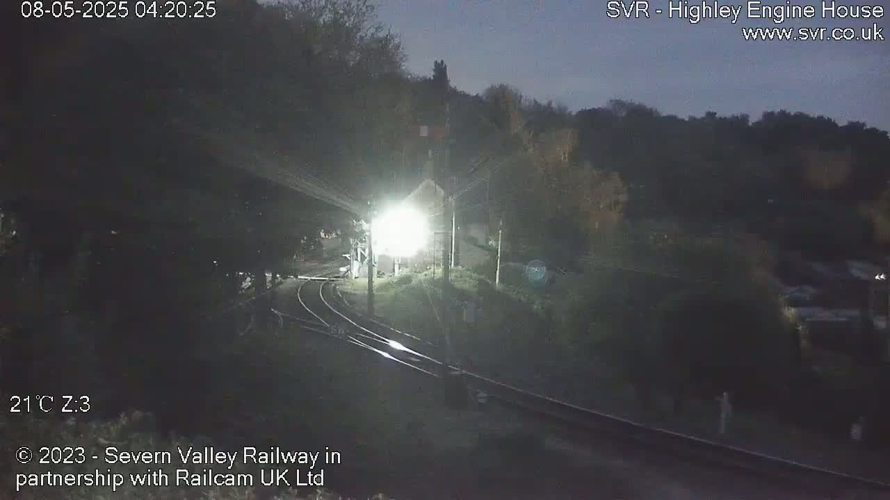A nighttime view of a brightly lit railway station surrounded by dark trees under a twilight sky.