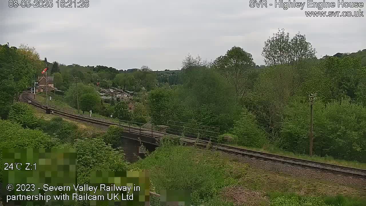 A railway track crosses a bridge over a small valley under an overcast sky, surrounded by lush green vegetation.