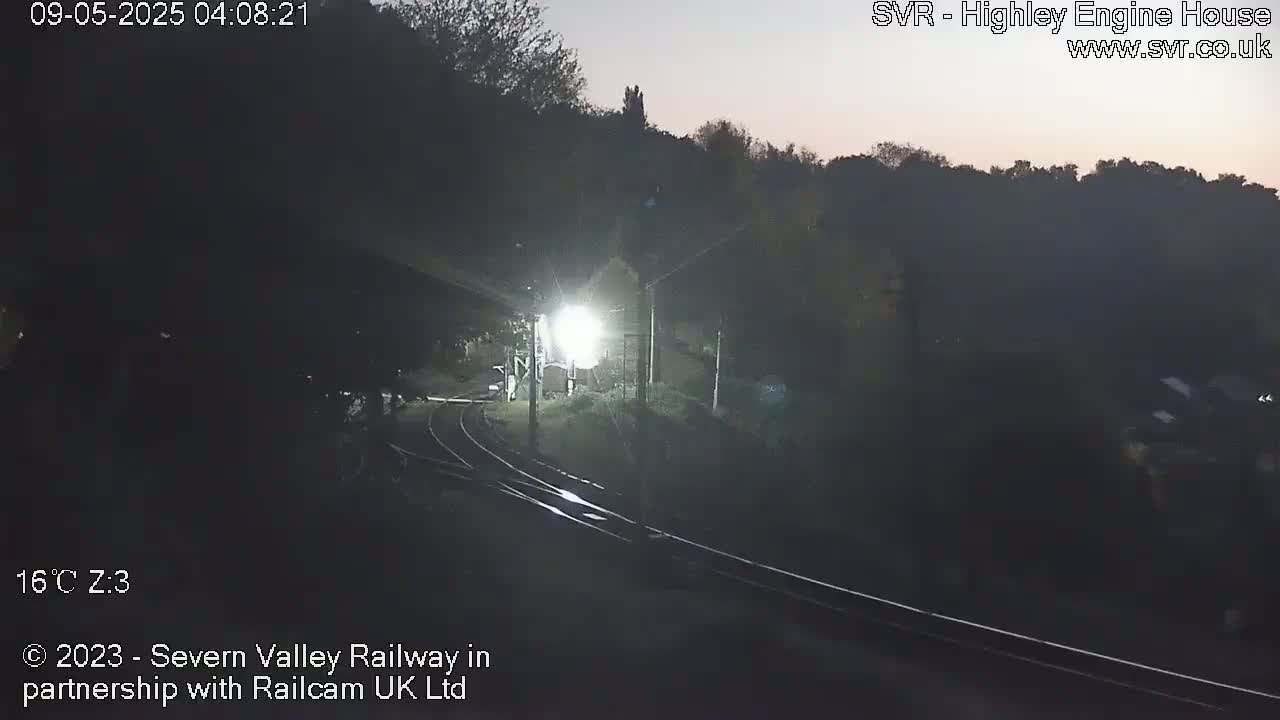 A nighttime, dimly lit view of a train station with train tracks and a bright light illuminating a portion of the area.