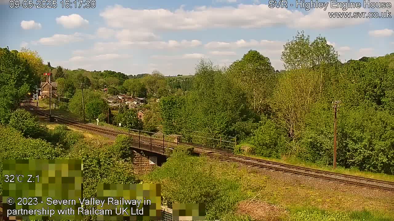 A sunny day reveals a railway track winding through a verdant landscape, passing over a low stone bridge near a small building, with lush green trees and shrubs lining both sides of the tracks.