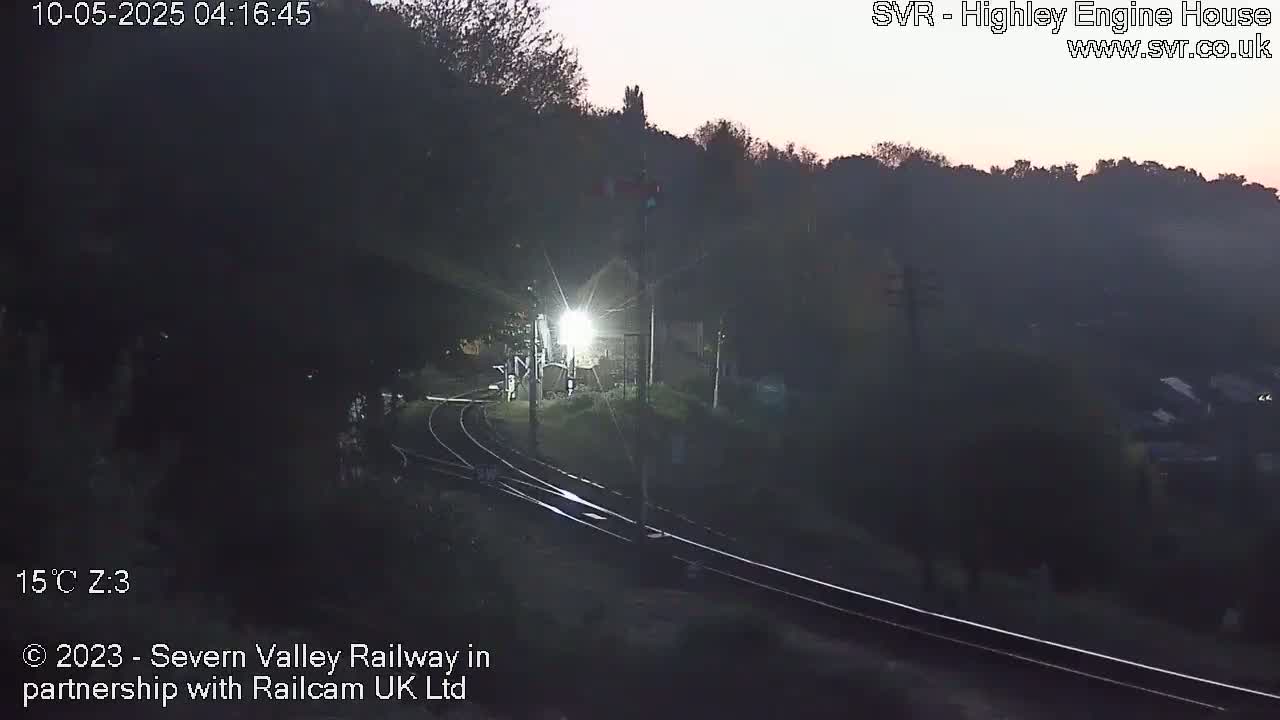 A brightly lit train approaches a dimly lit railway station at dawn, surrounded by dark hills and trees.