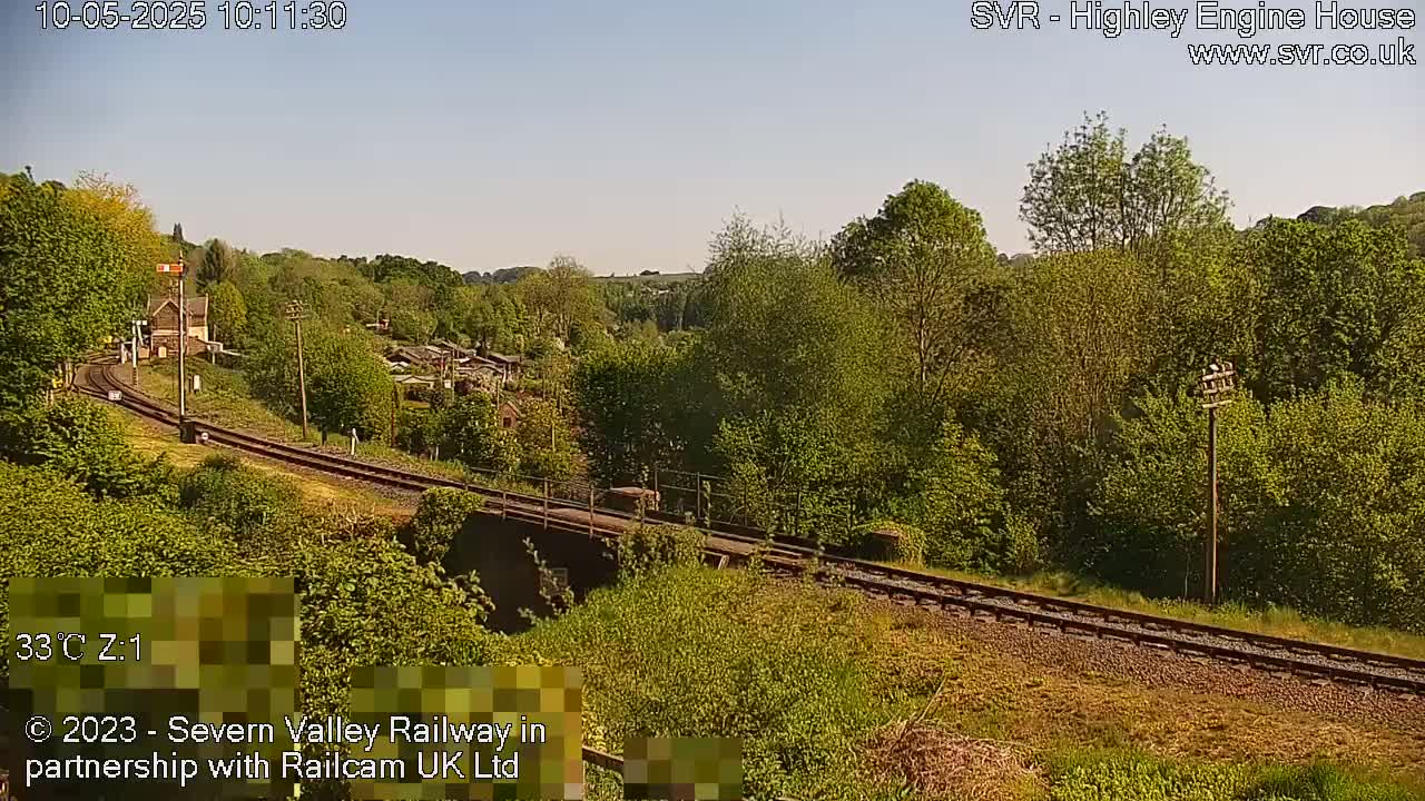 A sunny view of a railway track curving through a lush green landscape with trees and a small building visible in the distance.