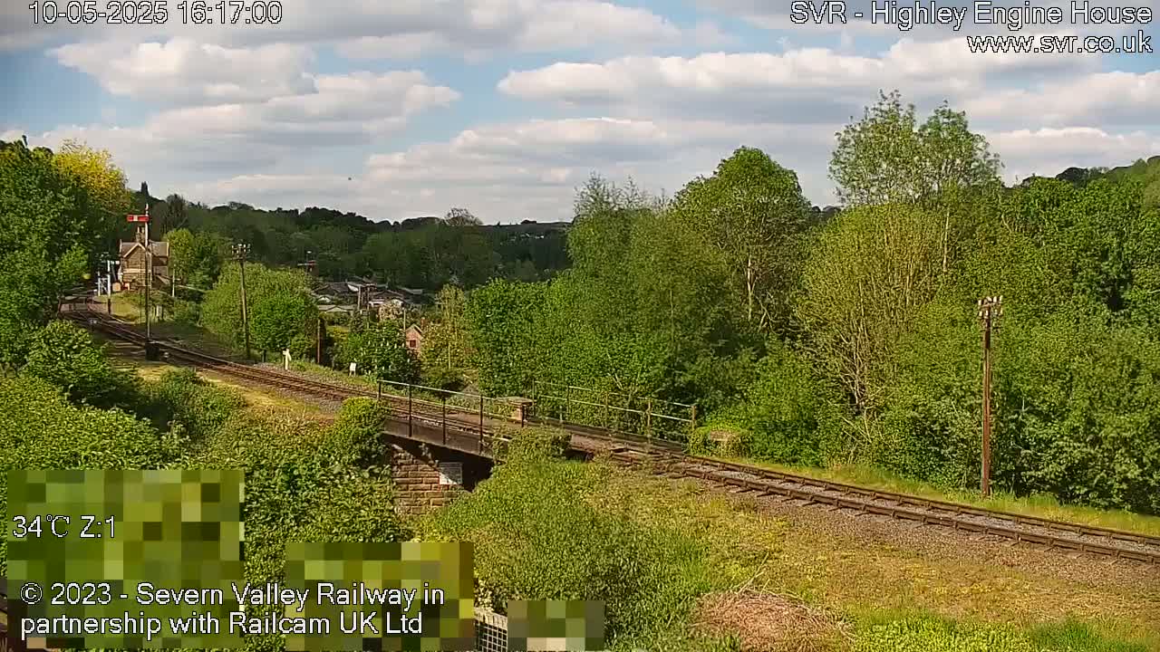 A partly cloudy day shows a railway track leading to a stone building nestled among lush green trees and vegetation.