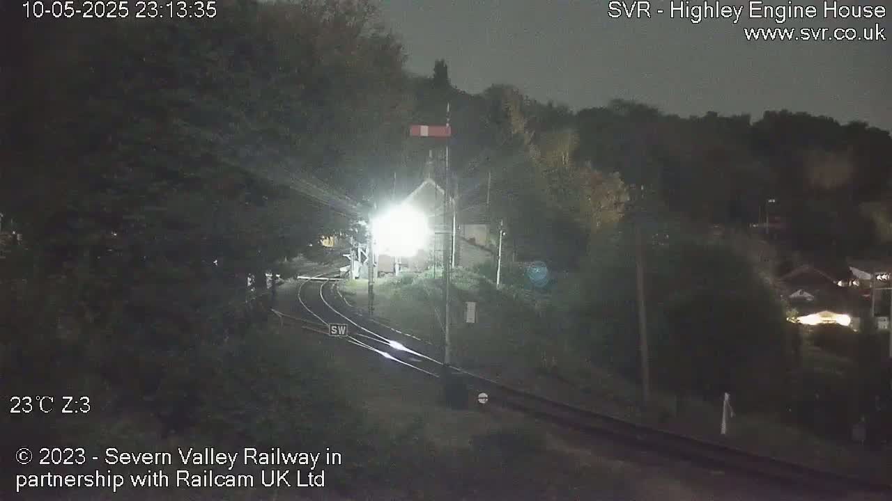 A nighttime view of a train approaching a station, surrounded by dark trees and illuminated by headlights.