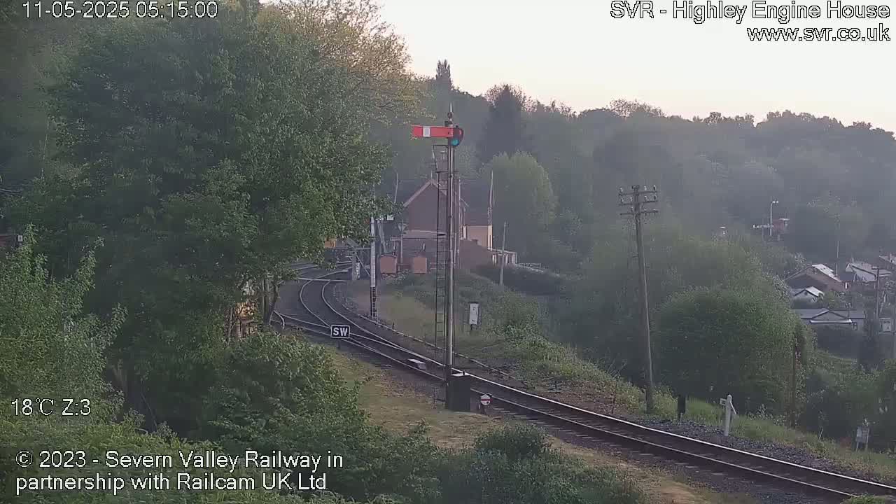 A hazy sunrise scene shows a railway track winding through a lush green hillside, with a signal and a small building visible in the mid-ground.
