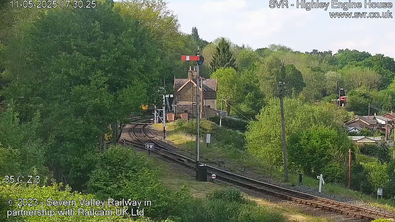 A train approaches a railway station nestled amongst lush green trees on a sunny day.