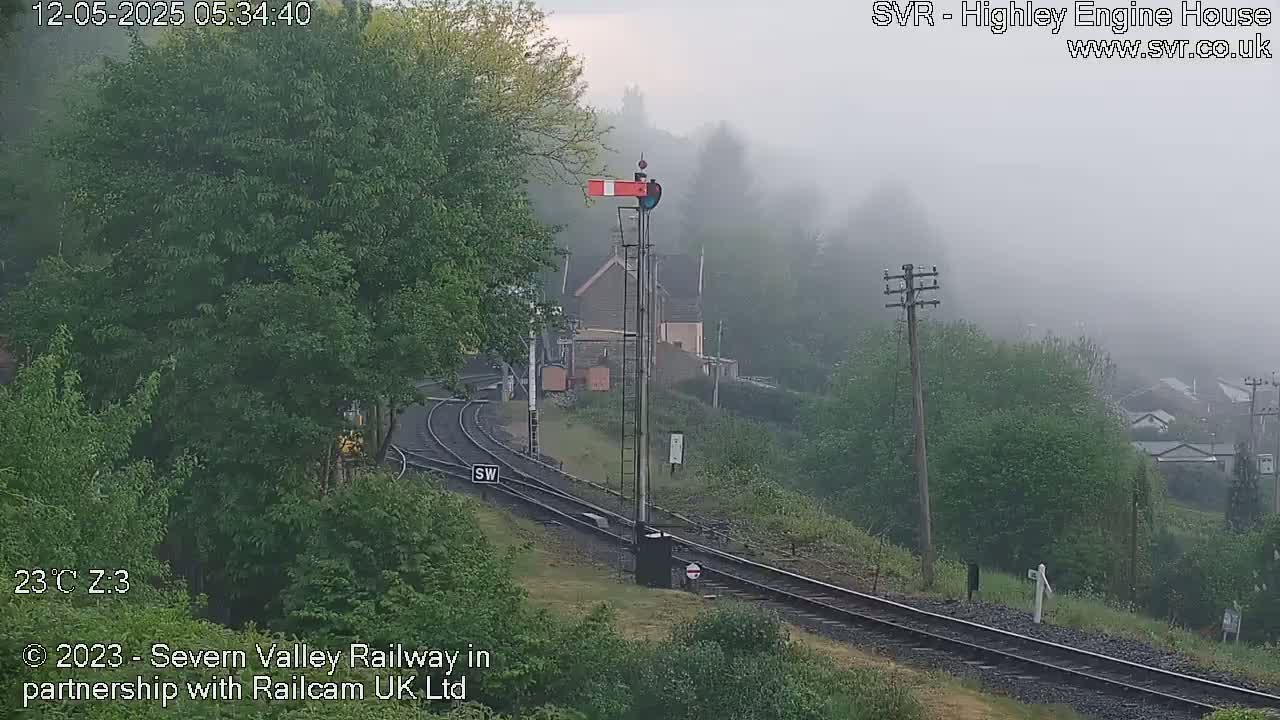 A foggy morning scene shows a railway track curving away from the camera, past a signal and a small building, toward a hillside shrouded in mist.