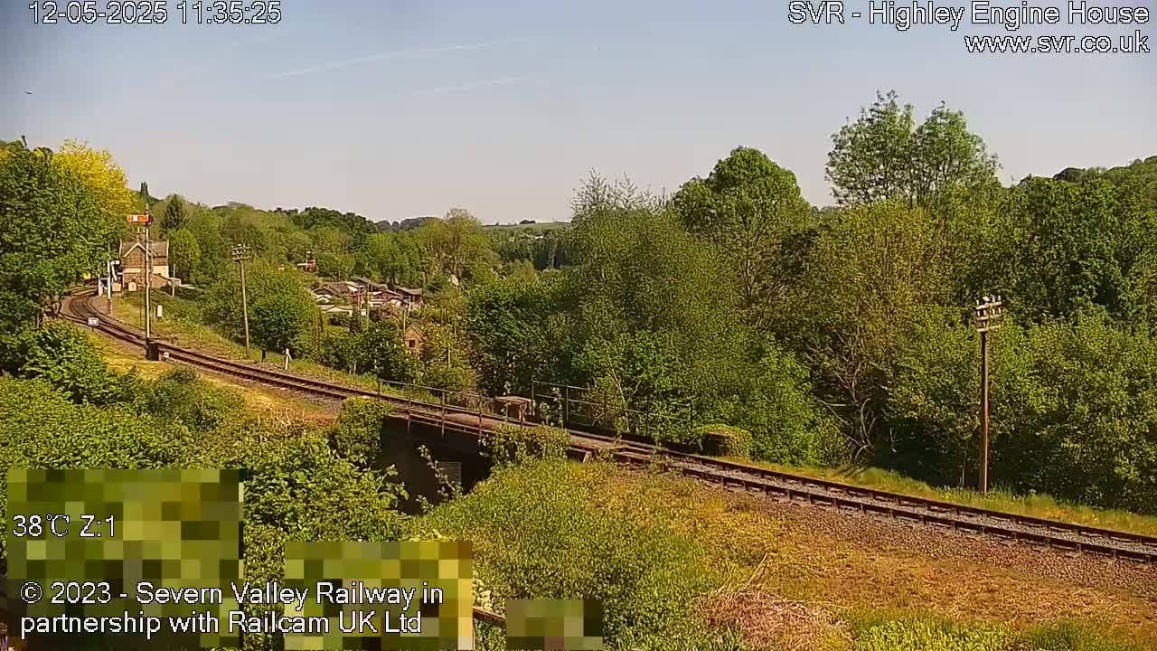 A sunny view of a train track curving through a lush green landscape, with a small building and houses visible in the distance.