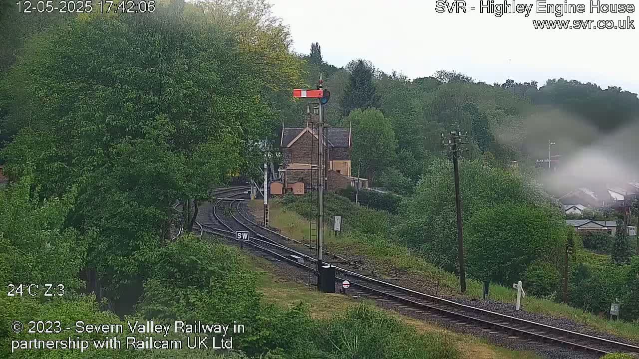 A train track curves through a lush green landscape, alongside a stone building and signal, under an overcast sky.