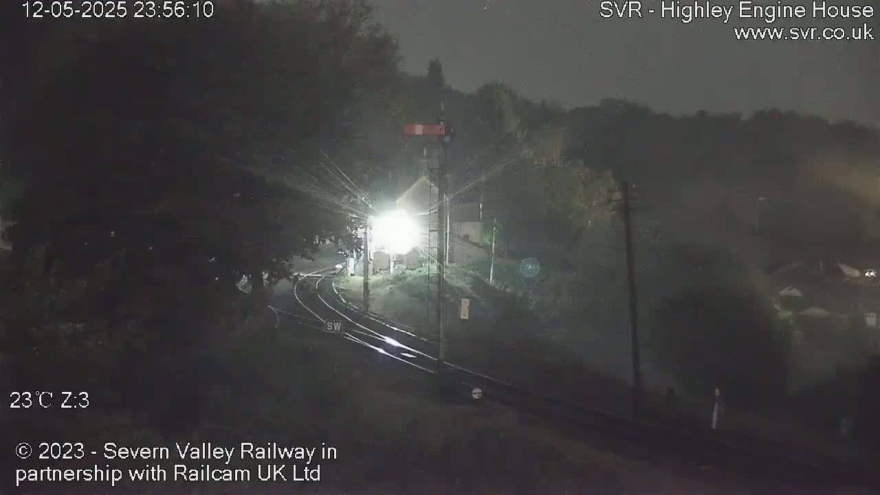 A nighttime view of a railway crossing with a signal light, illuminated by a nearby lamp, under a dark, clear sky.